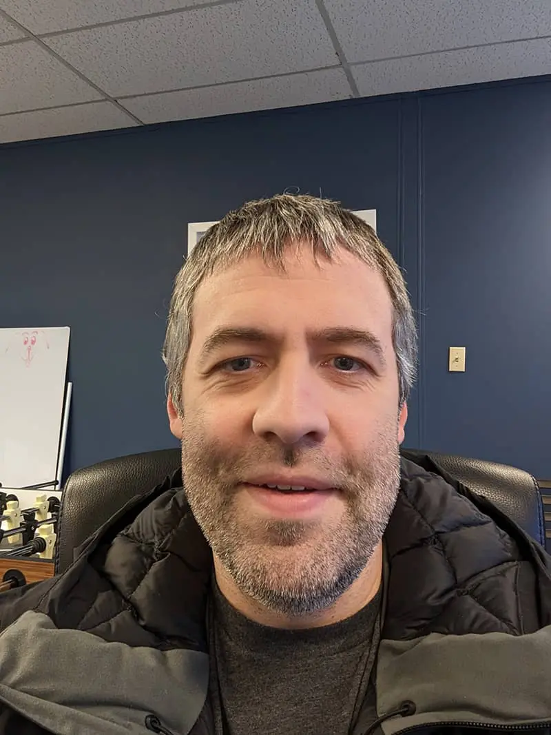 Man with light brown hair and beard wearing black puffy jacket in office with dark blue wall
