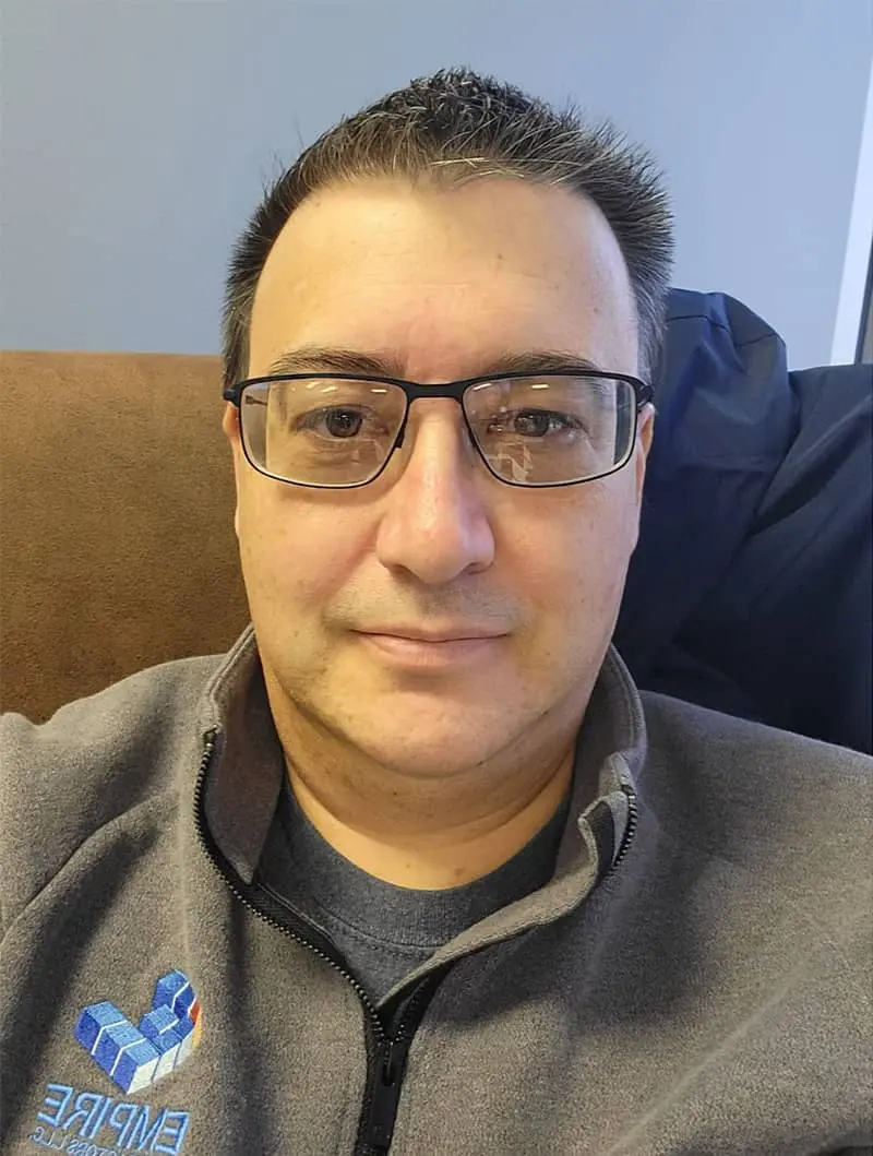 Man wearing glasses and tan jacket with blue logo, smiling at camera while seated indoors