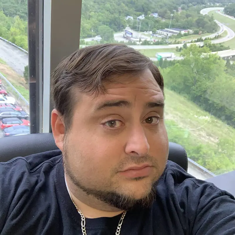 Man in dark shirt with silver chain necklace in high-rise office with expansive view of Cincinnati valley, parking lot, and green landscape below
