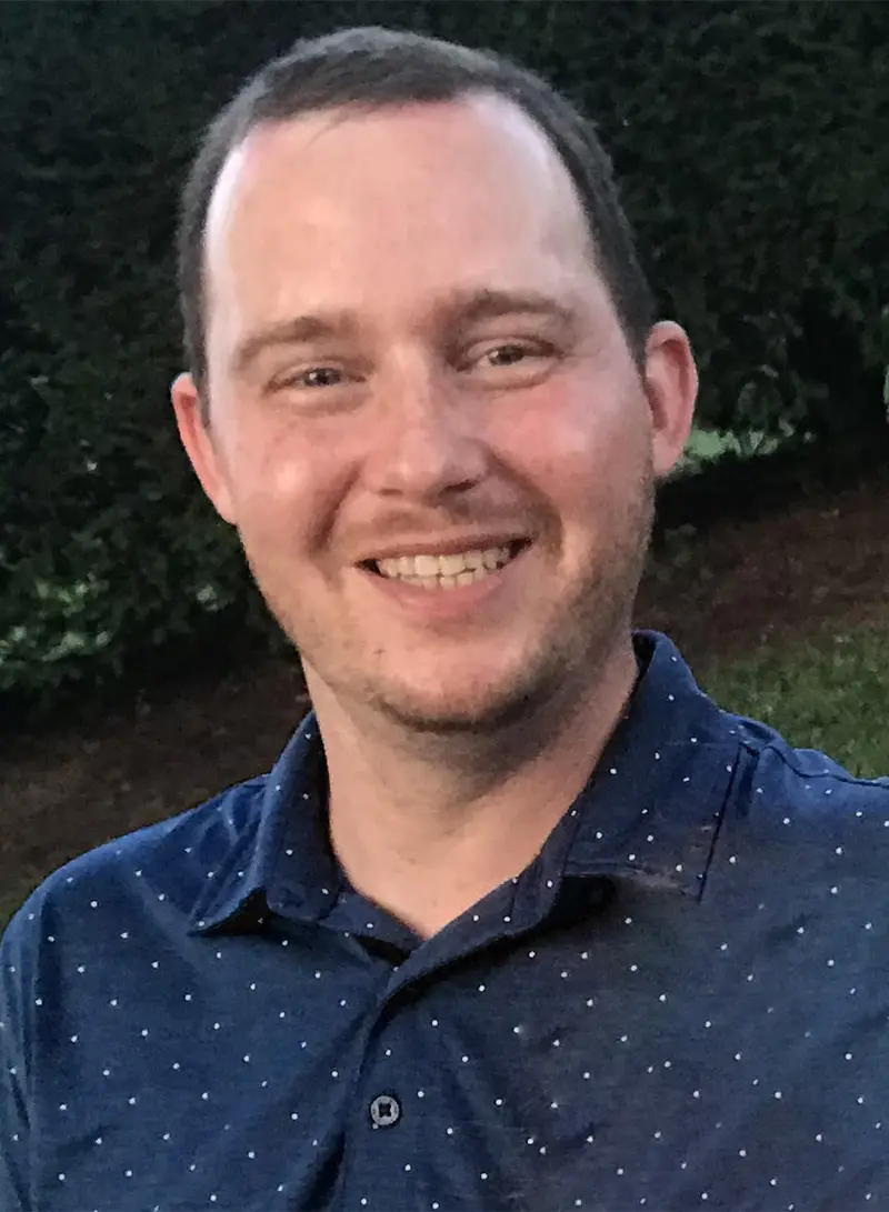 Smiling man wearing a dark blue button-up shirt with white speckles, photographed outdoors against a green hedge background
