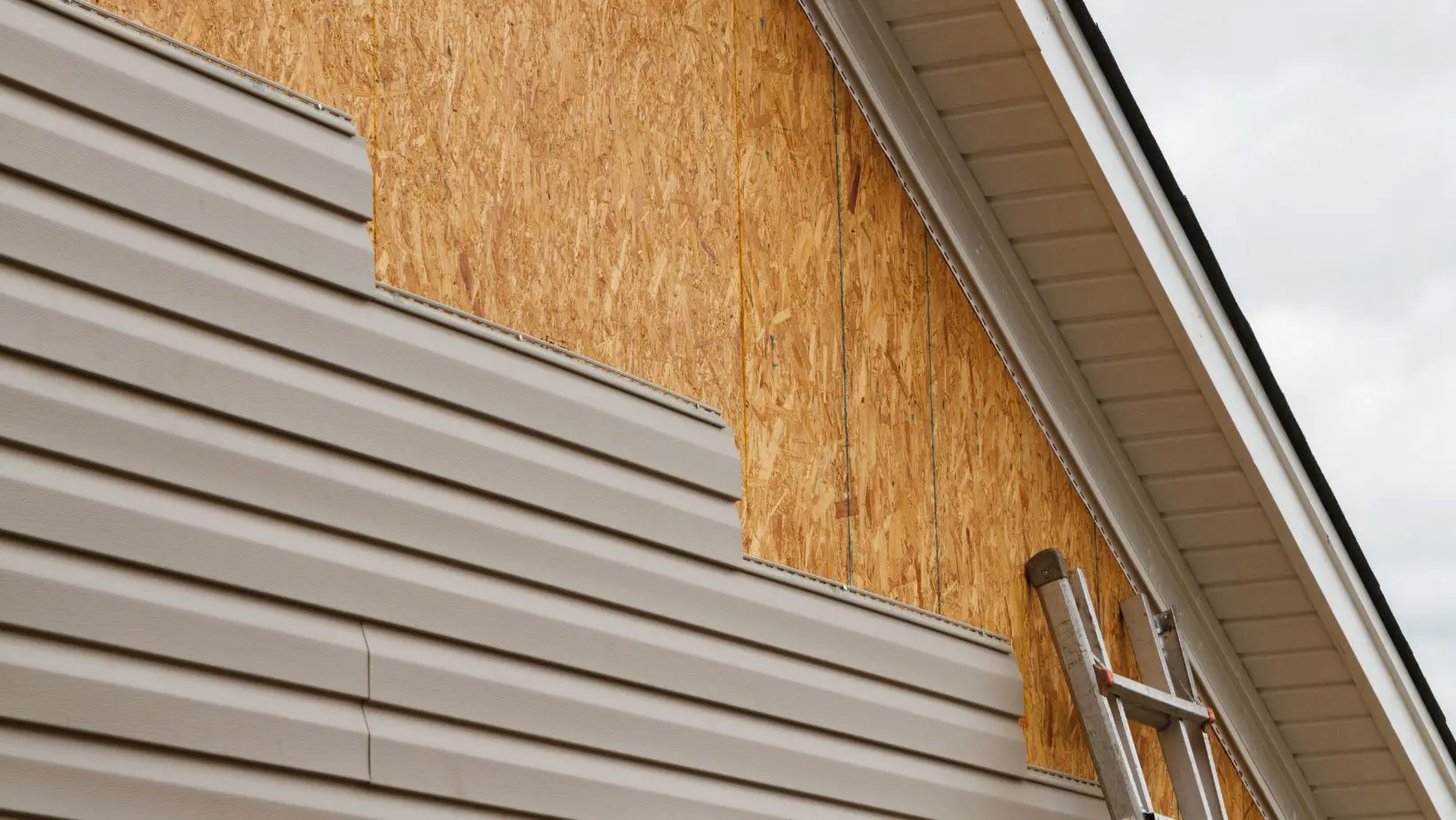 Partially installed beige vinyl siding on a home exterior, revealing the wooden sheathing underneath. A ladder is positioned for ongoing construction or repair. Empire Contractors provides professional siding installation and replacement for durable, weather-resistant home exteriors.