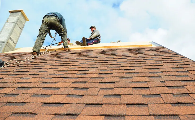 Two roofers installing asphalt shingles on a residential roof with a chimney and roof vent visible under clear skies