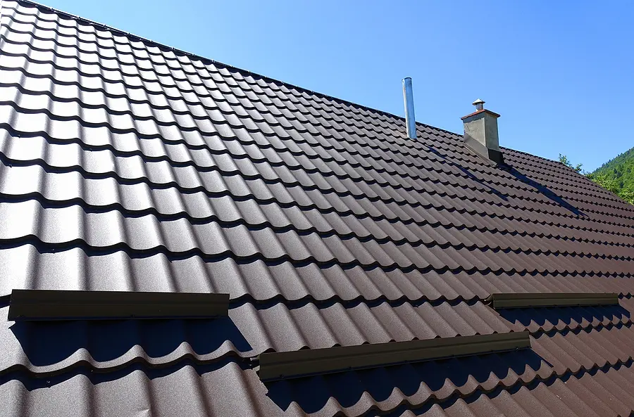 Modern brown metal profile roof with corrugated wavy shingles, chimney flashing, and roof vent on residential house exterior