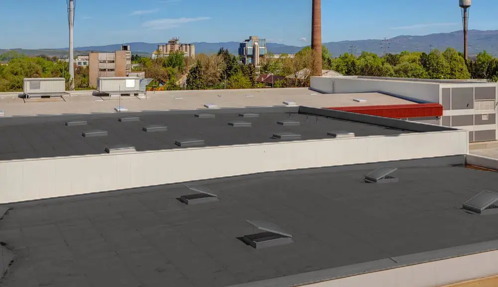 Flat commercial roofing with black membrane, roof vents, and white parapet walls overlooking Cincinnati skyline with mountains