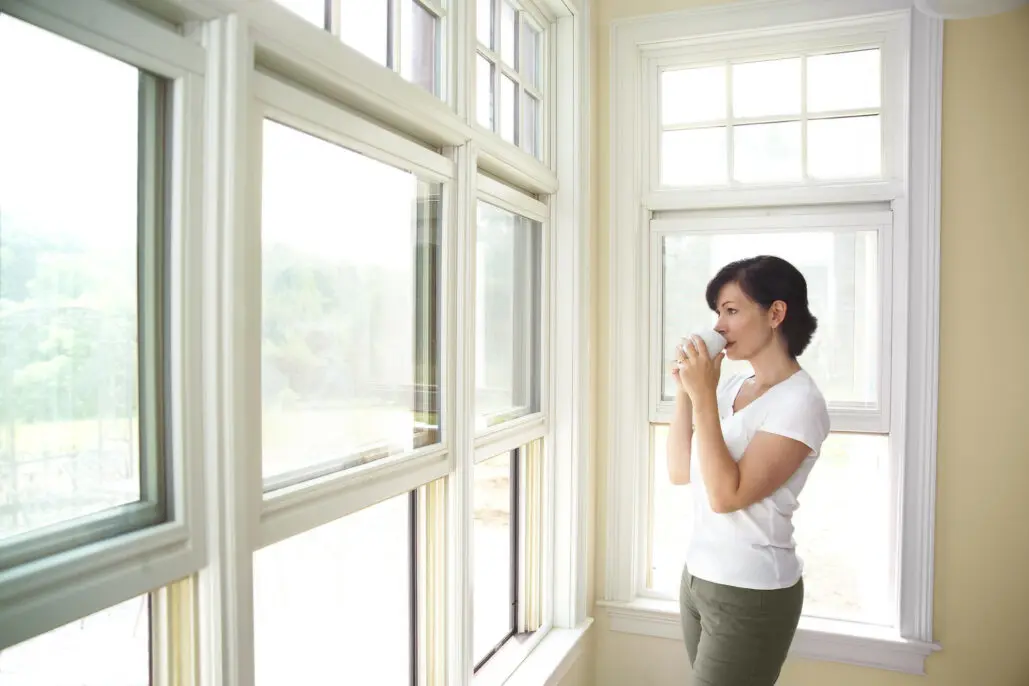Woman looking out through large white vinyl replacement windows in a bright room with scenic outdoor view