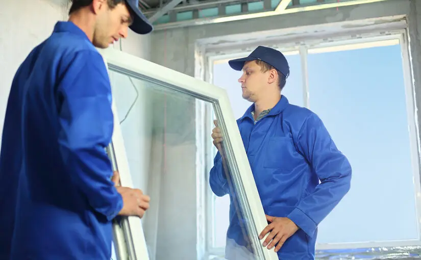 Two workers in blue uniforms and caps installing a vinyl window frame in an unfinished room with bright natural light