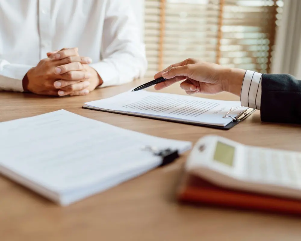 Two business professionals at a wooden desk reviewing a document with a pen, papers, and calculator visible during a consultation meeting