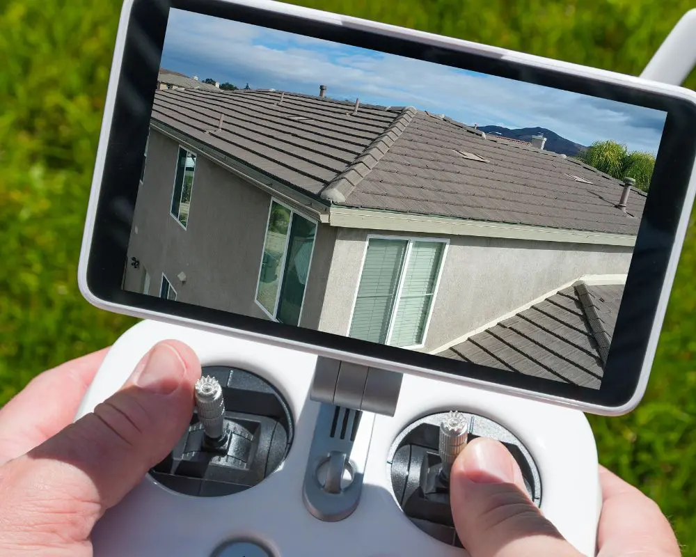 Drone controller displaying aerial view of residential home with dark asphalt shingle roof, gray siding, and multiple windows against green lawn background