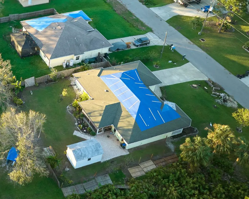 Aerial view of residential properties with blue tarped roofs indicating storm damage repair work, showing asphalt shingle roofing and residential siding in a suburban neighborhood