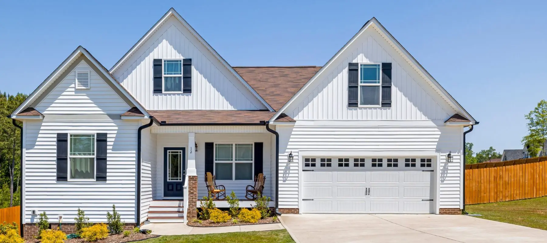 Modern white farmhouse with black shutters, brick chimney, two-car garage, and manicured landscaping under clear blue sky