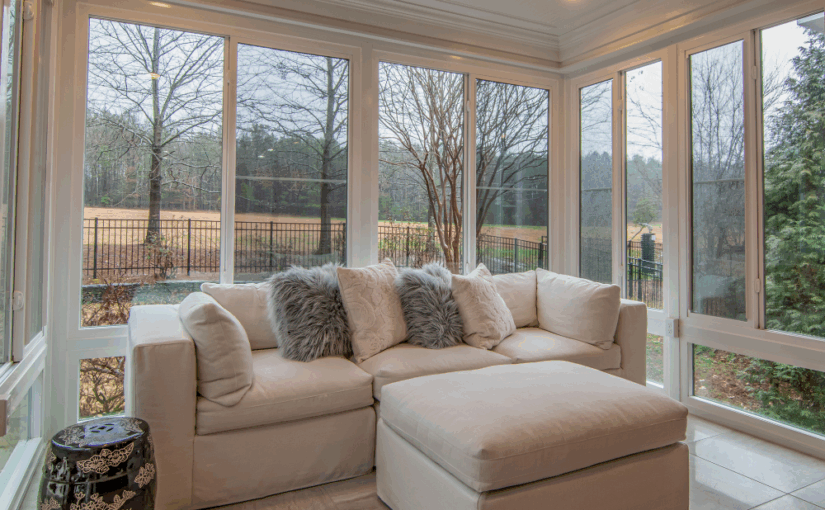 Sunroom with cream sectional sofa, decorative pillows, and ottoman overlooking bare trees and yard through expansive windows with white trim and ceiling fan