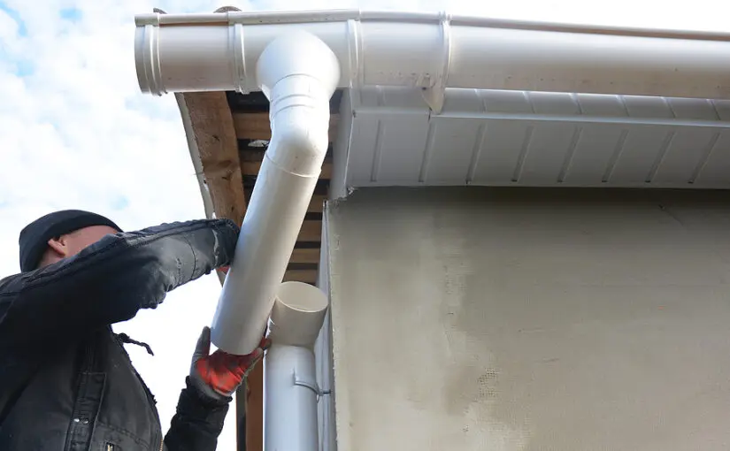 Worker installing white gutter system and downspout on residential roof edge against cloudy sky