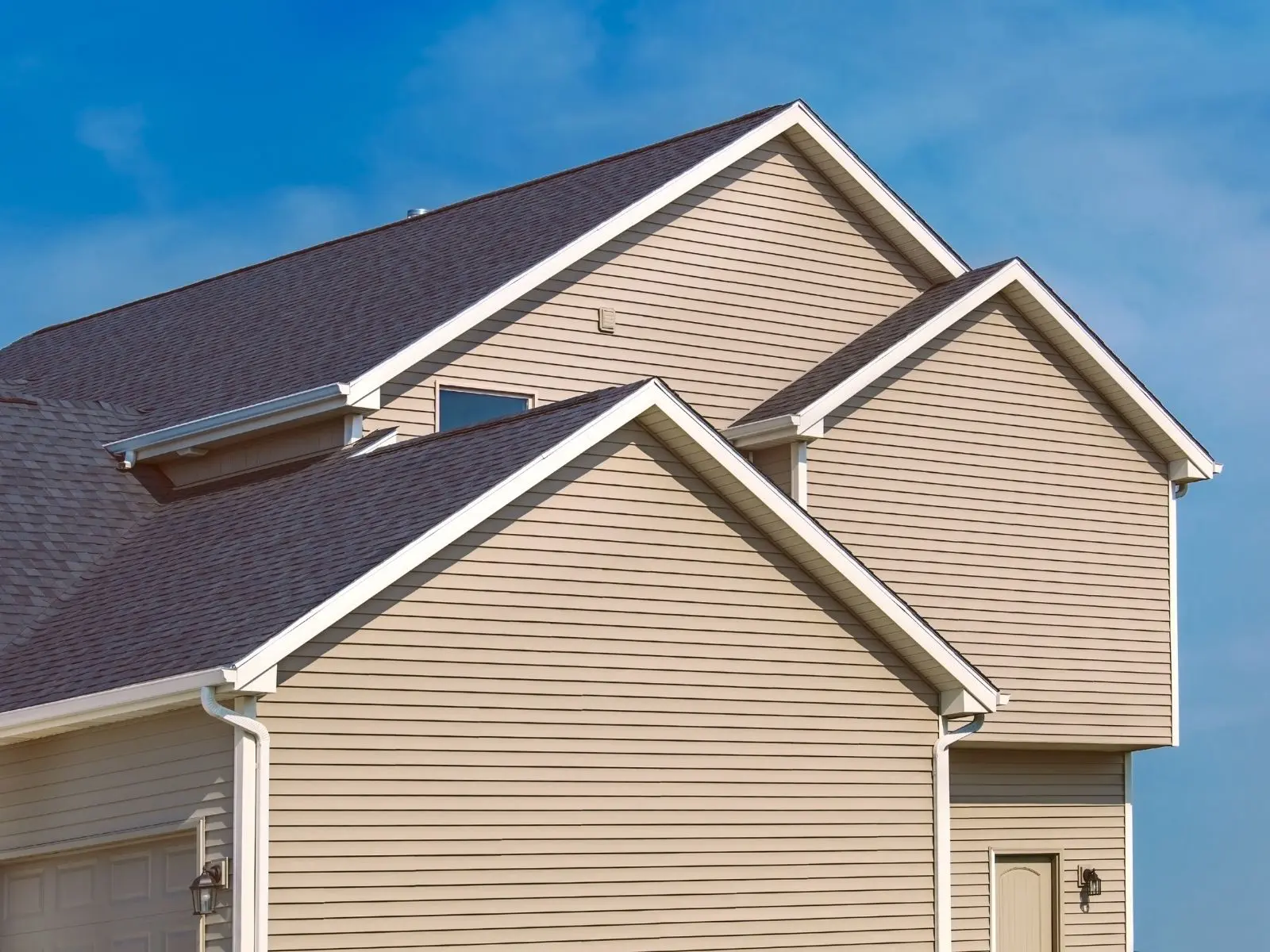 Modern home with beige vinyl siding, dark asphalt shingle roof, multiple gables, and attached garage under clear blue sky