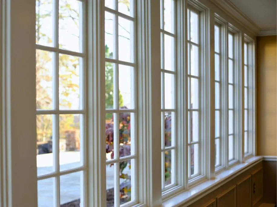Sunlit interior view of multiple tall vinyl windows with grid panes, overlooking a garden with autumn foliage and flowering plants