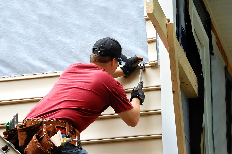 Worker in red shirt installing vinyl siding on house exterior wall during home repair project