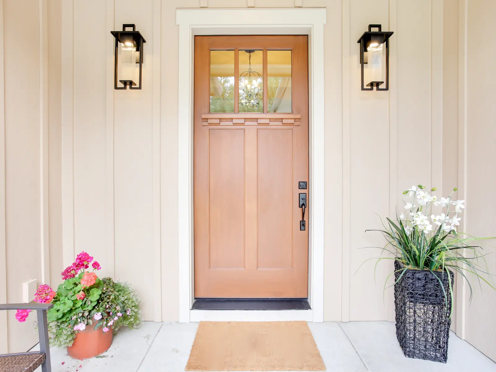 Modern peach-colored entry door with three glass panes set in white trim, flanked by black wall sconces and flowering planters on a light-colored porch