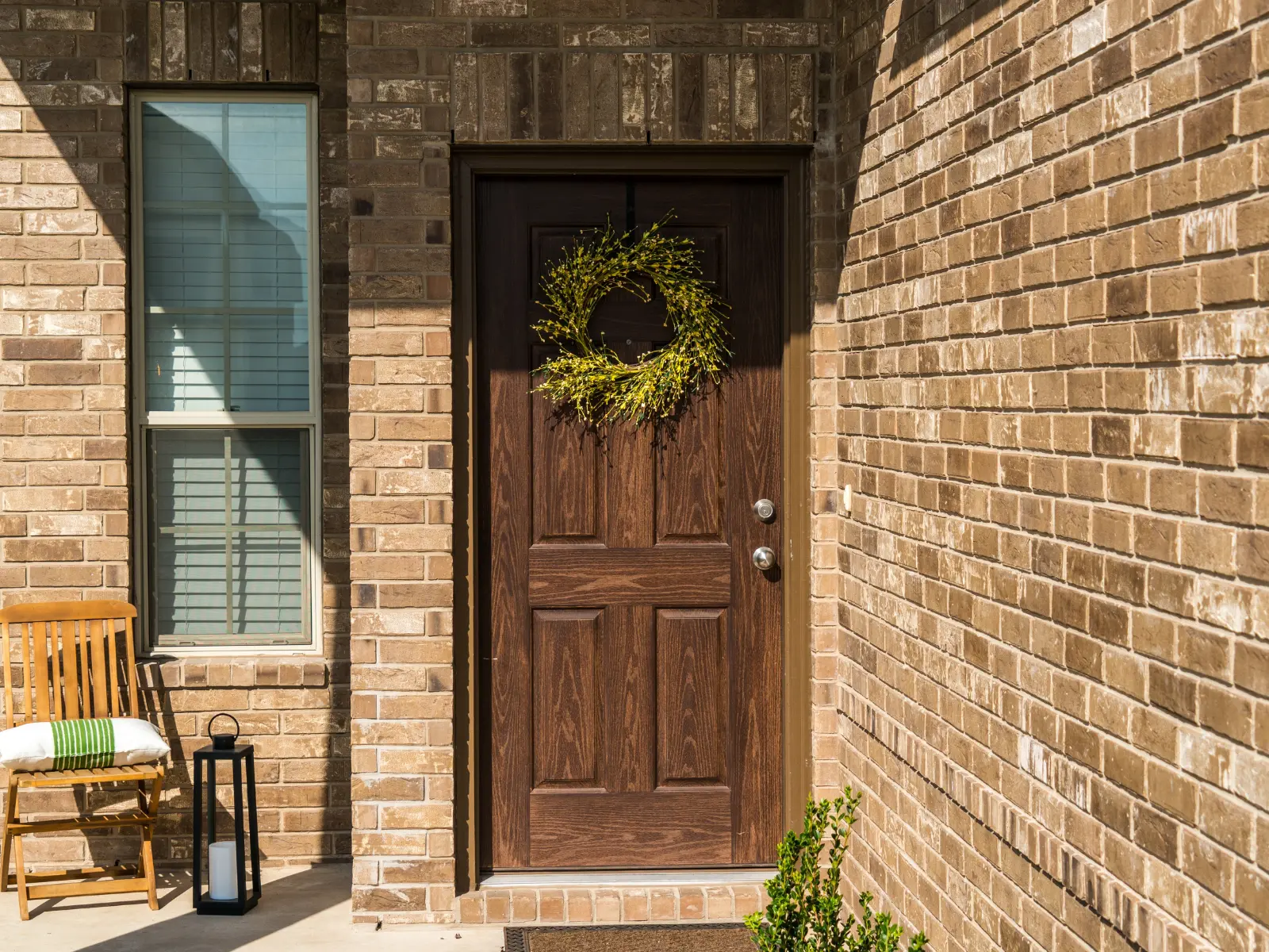 Brick home entryway with dark wood entry door decorated with yellow wreath, window with shutters, wooden chair, and black lantern on porch