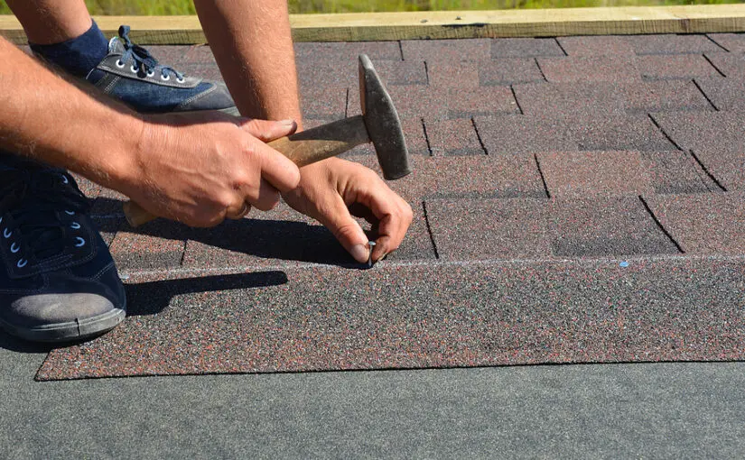 Roofer using hammer and nail to install asphalt shingles on residential roof during repair or replacement project