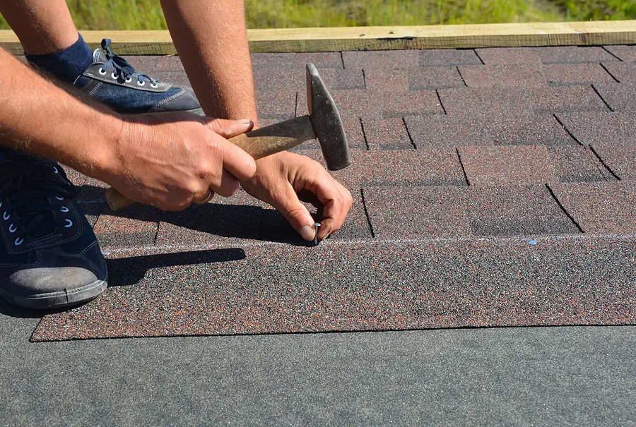 Roofer using hammer and nail to install asphalt shingles on residential roof during repair or replacement project