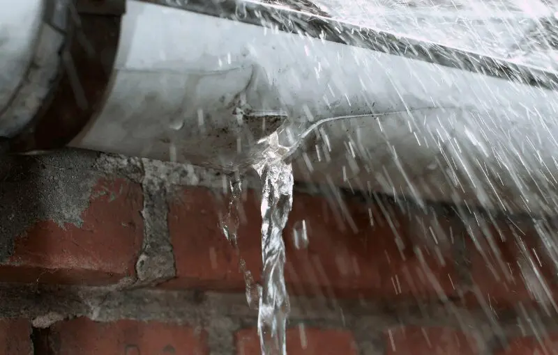 Water streaming from a roof gutter during heavy rain, showing roof flashing and brick siding below, illustrating potential roof leak and gutter issues