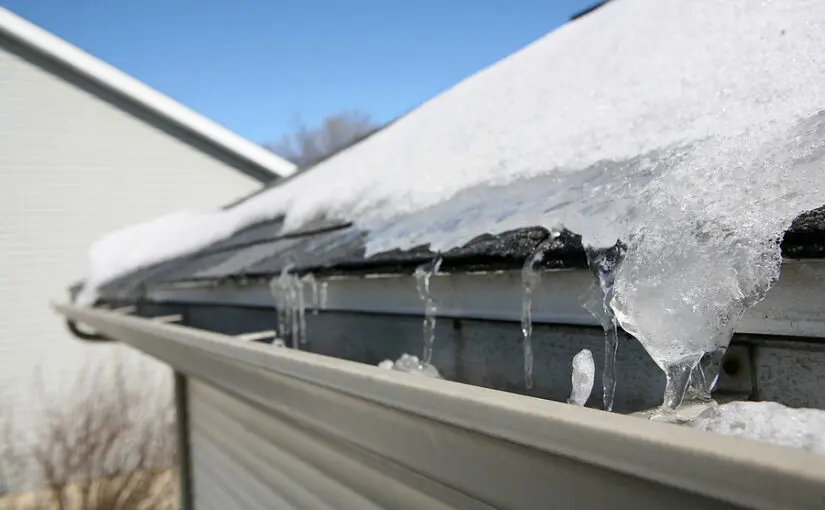 Icicles and ice buildup on roof gutters and shingles with snow-covered roof in background on clear winter day