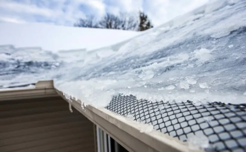Icy buildup and snow accumulation on residential roof with mesh gutter guard and winter landscape background, showing ice dam formation and potential roof damage