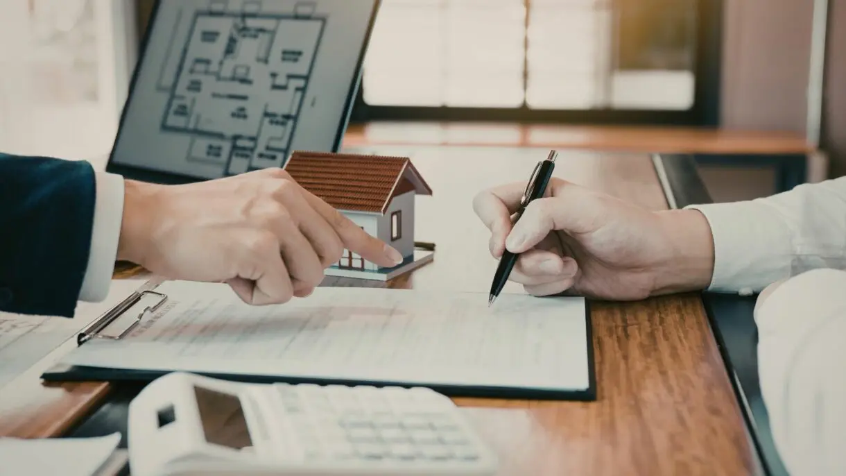 Real estate agent and client reviewing property documents at desk with architectural model house and floor plans