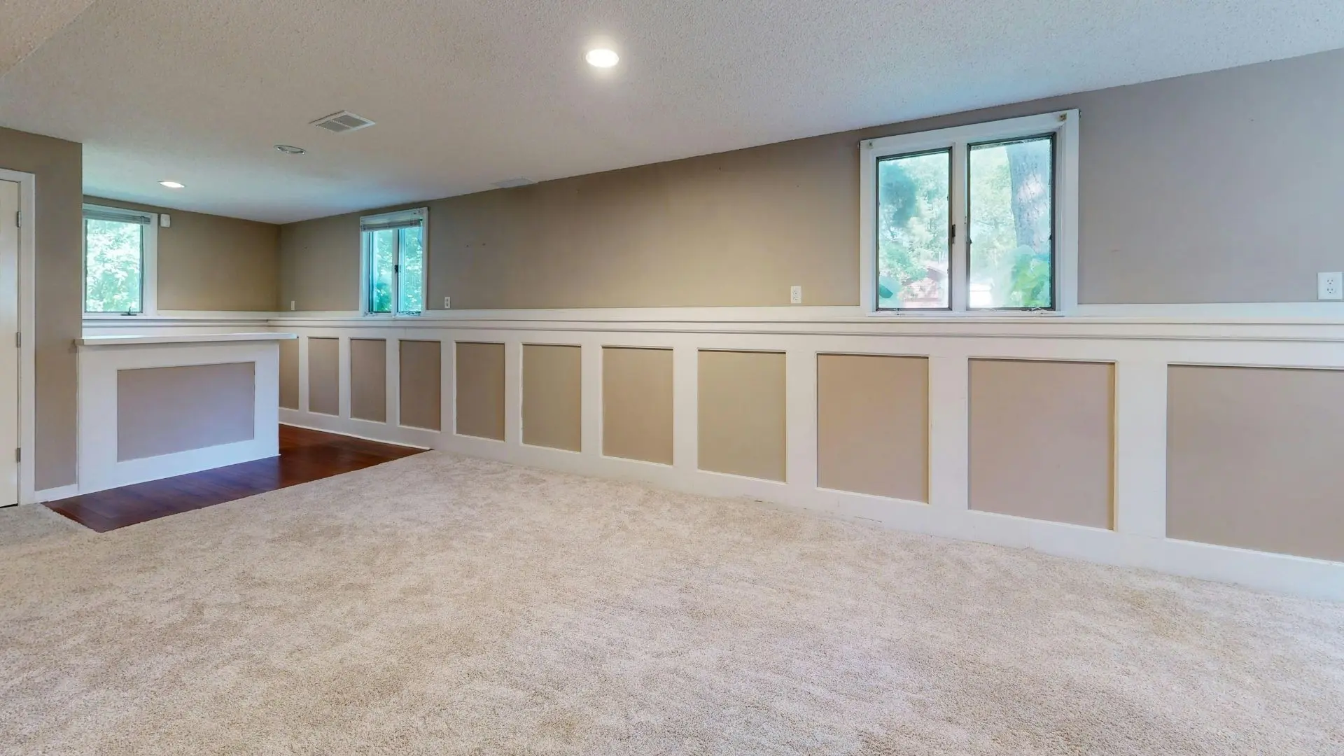 Spacious basement room with beige walls, white wainscoting panels, carpet flooring, large windows, recessed lighting, and a fireplace accent wall
