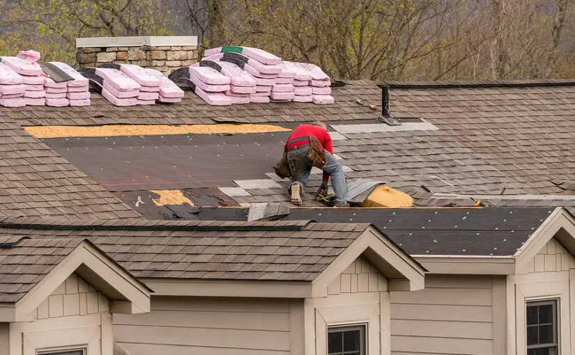 Roofing contractor removing old shingles from a residential roof during replacement, with pink shingle bundles and tools visible on the upper roof surface