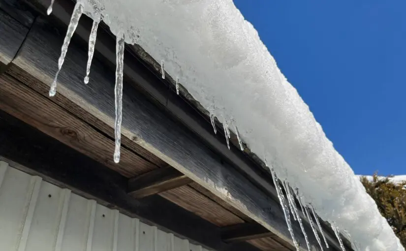 Icicles hanging from a frost-covered roof edge and wooden soffit against a clear blue sky, showing winter ice buildup and potential ice dam conditions