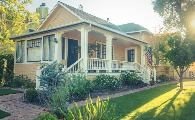 Charming cream-colored cottage with covered porch, white columns, asphalt shingle roof, manicured lawn, and mature trees in Cincinnati neighborhood