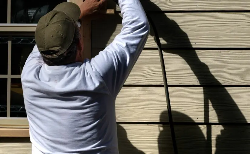 Worker in white shirt and tan cap using measuring tape on wooden siding during roof and exterior home inspection