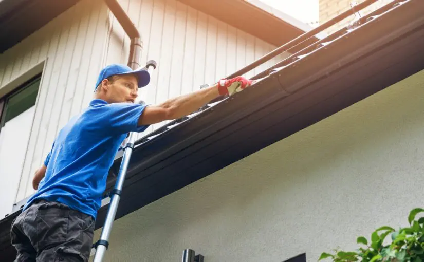 Worker in blue shirt and cap on ladder inspecting or cleaning gutters on residential roof and fascia