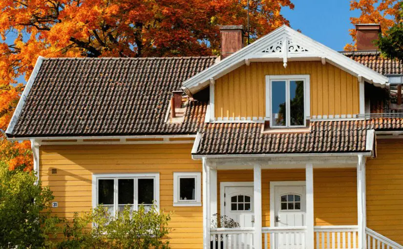 Yellow wood-sided two-story house with white trim, tile roof, front porch, and autumn trees with orange foliage in background