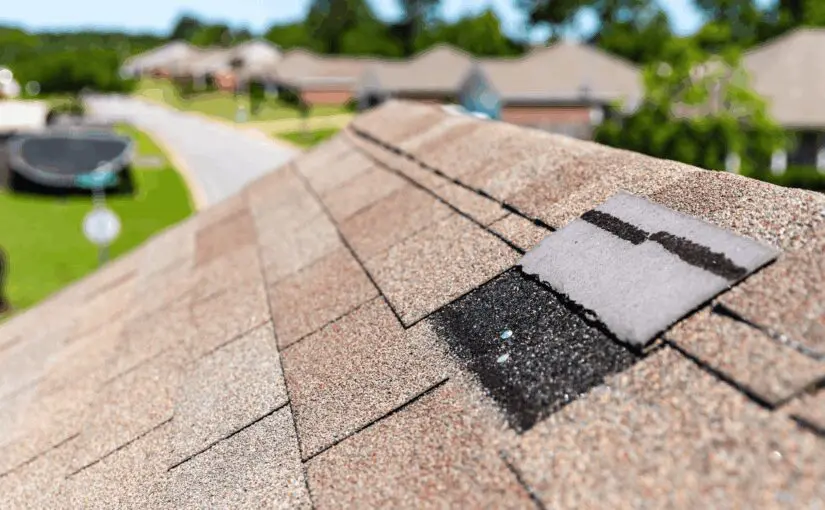 Close-up of asphalt shingle roof showing missing shingles and storm damage with residential neighborhood in background