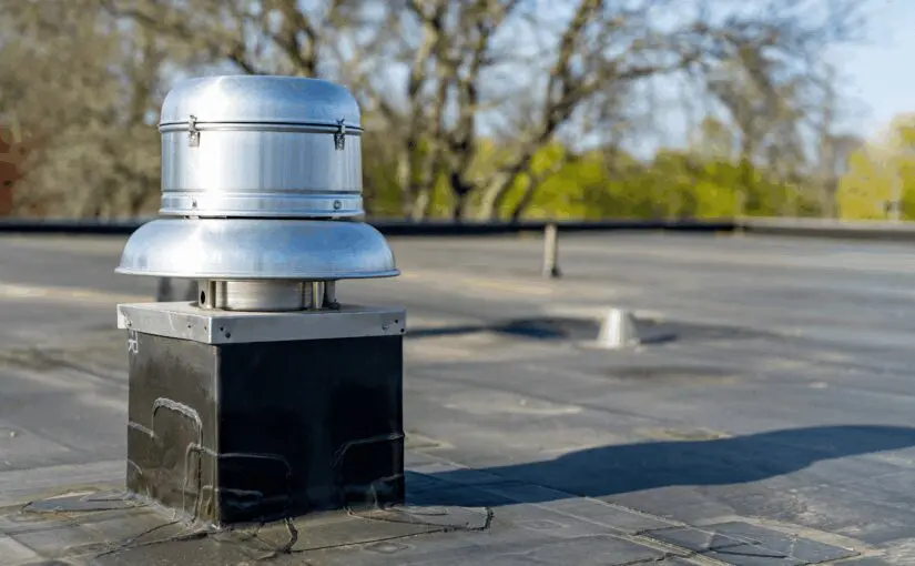 Stainless steel roof vent with dome cap mounted on flat commercial roofing, overlooking parking lot with trees