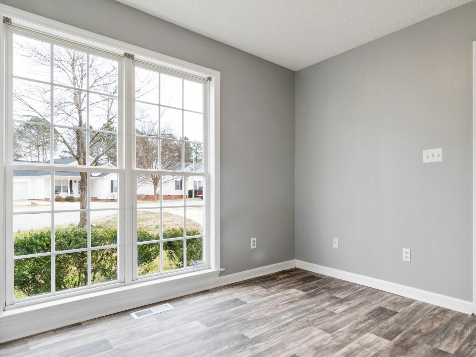 Empty bedroom with large multi-pane windows overlooking suburban street, gray walls, wood-look flooring, and white trim