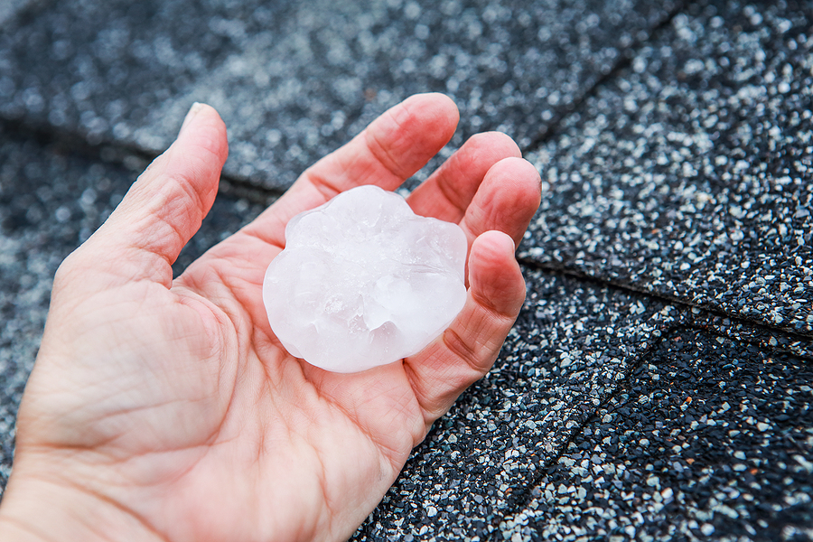 Hand holding a large piece of hail over asphalt roof shingles, showing potential impact damage that can weaken roofing materials.